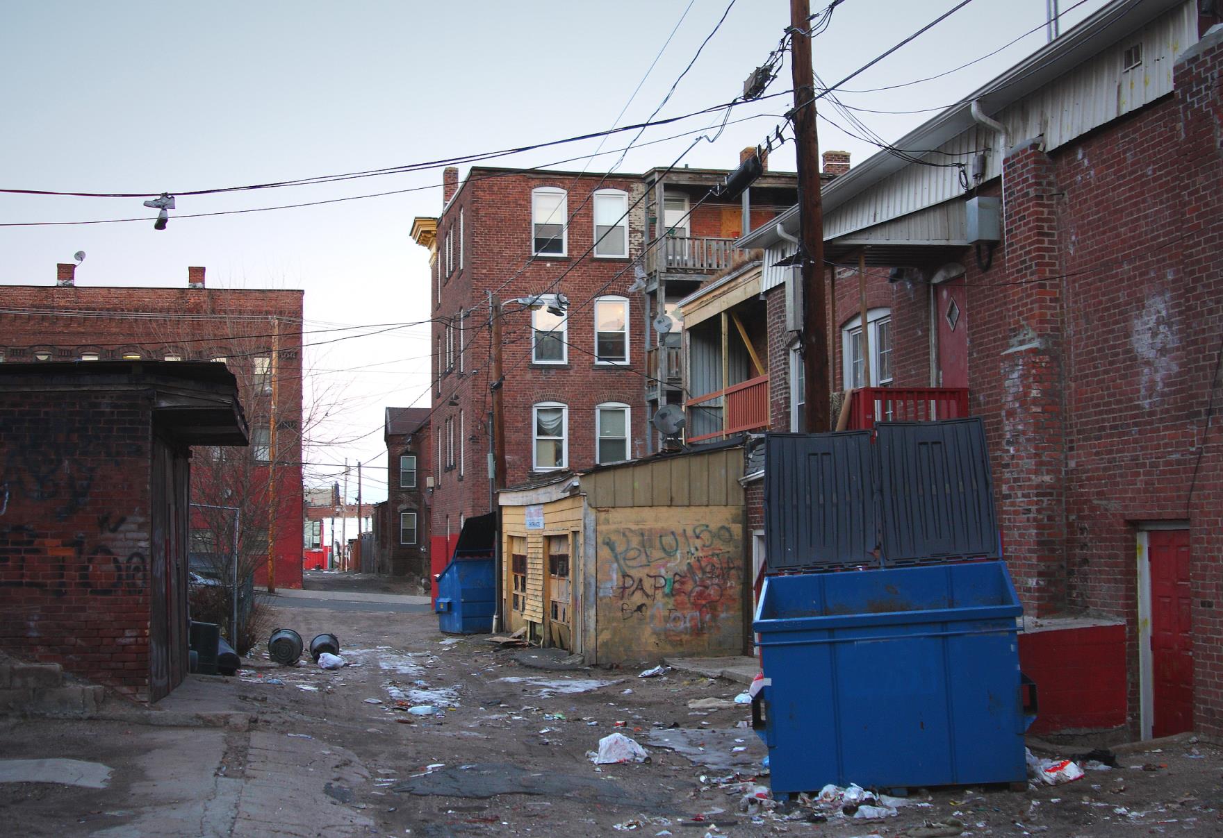 A trash-filled urban alleyway in an inner city urban neighborhood, with graffiti, trash and poorly maintained, aging buildings in the background.