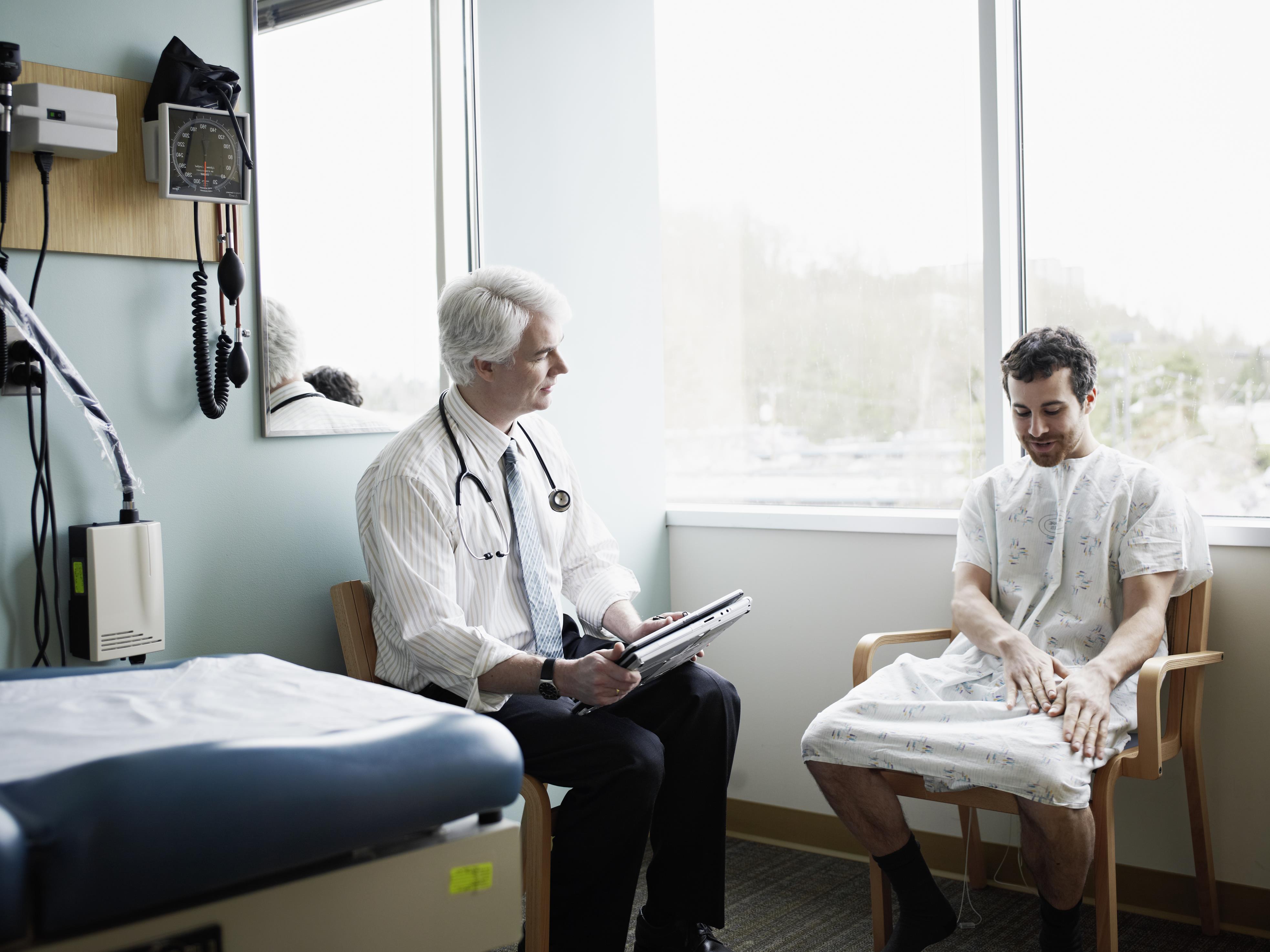 An older, white-haired, white doctor with a stethoscope hanging around his neck sits near a very young, dark-haired white man wearing as hospital gown and looking at his hands as he talks, as if he's a little embarrassed, a little nervous and wo<em></em>ndering why he feels a draft.