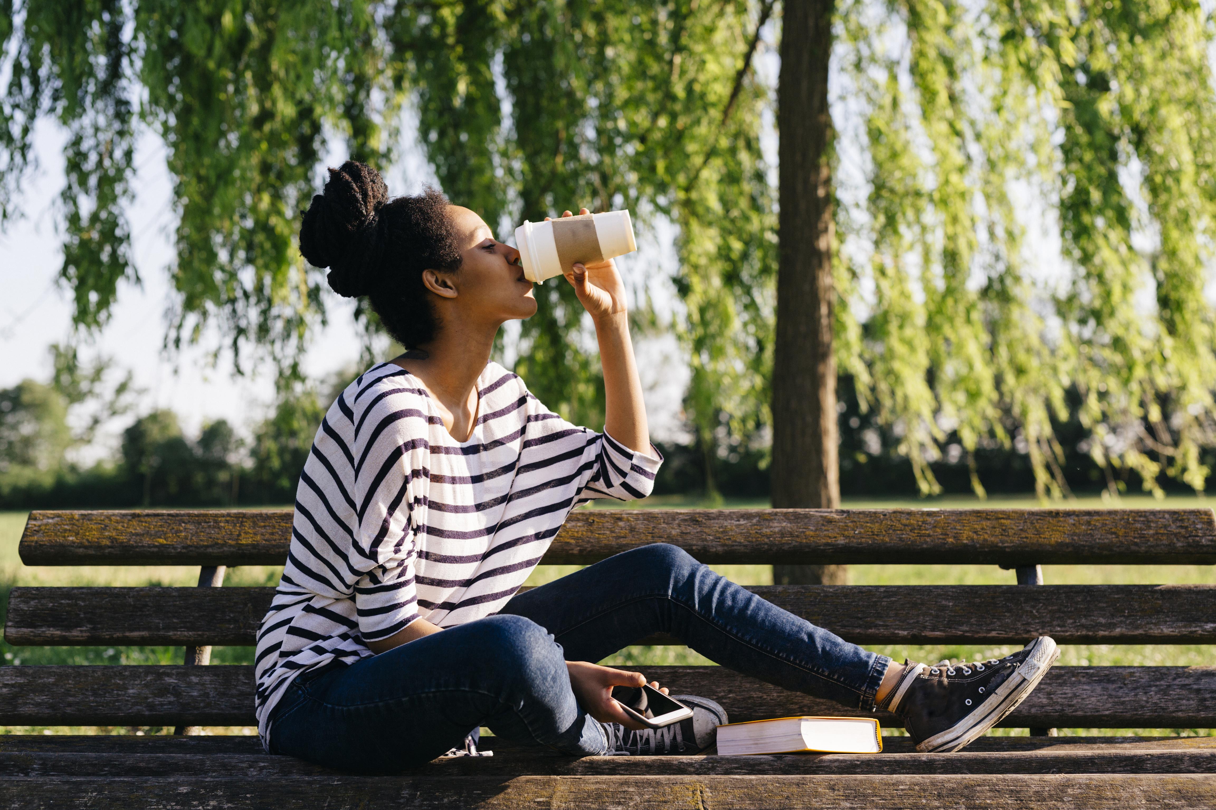 A young woman wearing casual apparel sitting on a park bench drinking coffee from a to-go cup.