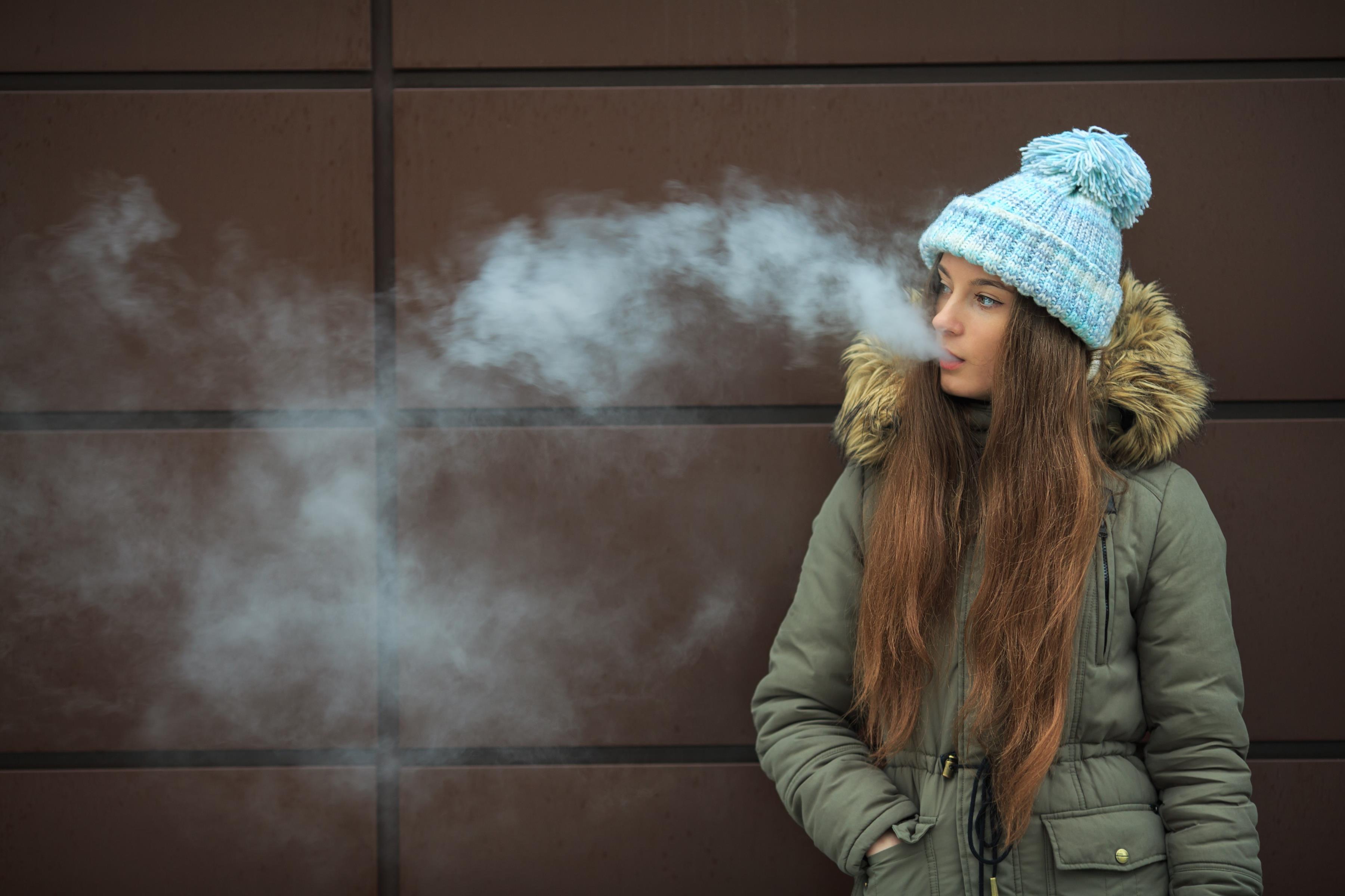 Wearing an overcoat and wool cap on a cold day, a teenage girl standing on the street smokes an electro<em></em>nic cigarette.