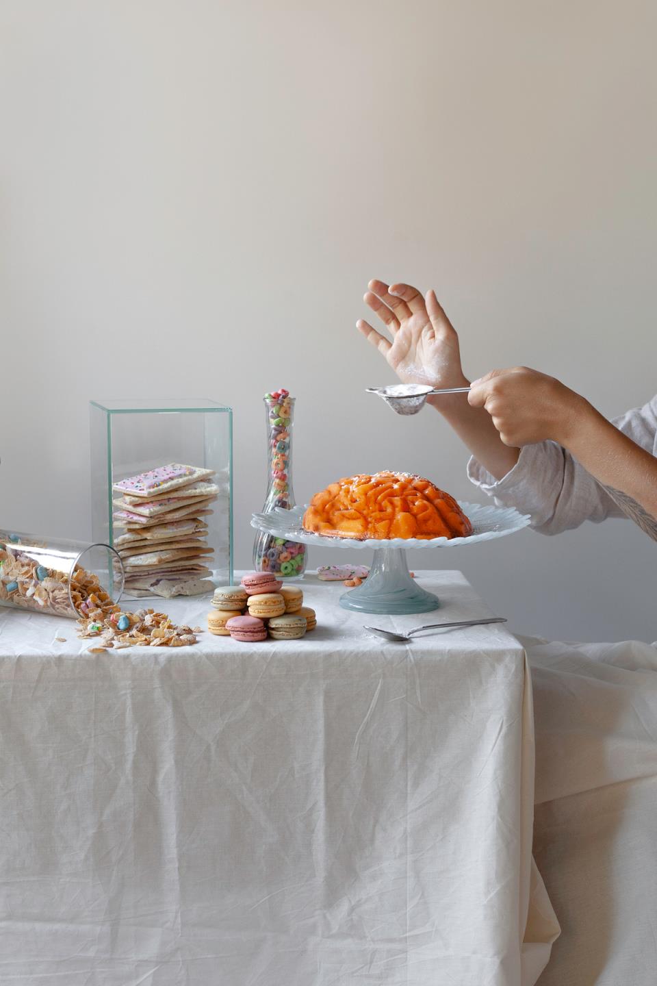 A jello mold in the shape of a brain sits on a platter, surrounded by sweets, while a person in the background holds a spoo<em></em>nful of sugar above the mold.